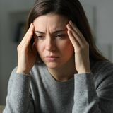 A woman with long brown hair wearing a gray knit sweater sits in a dimly lit room, holding both hands to her temples in a gesture of headache before using Solace™ Pain Support Formula - Flute Refill Nectar - BlissThera™
